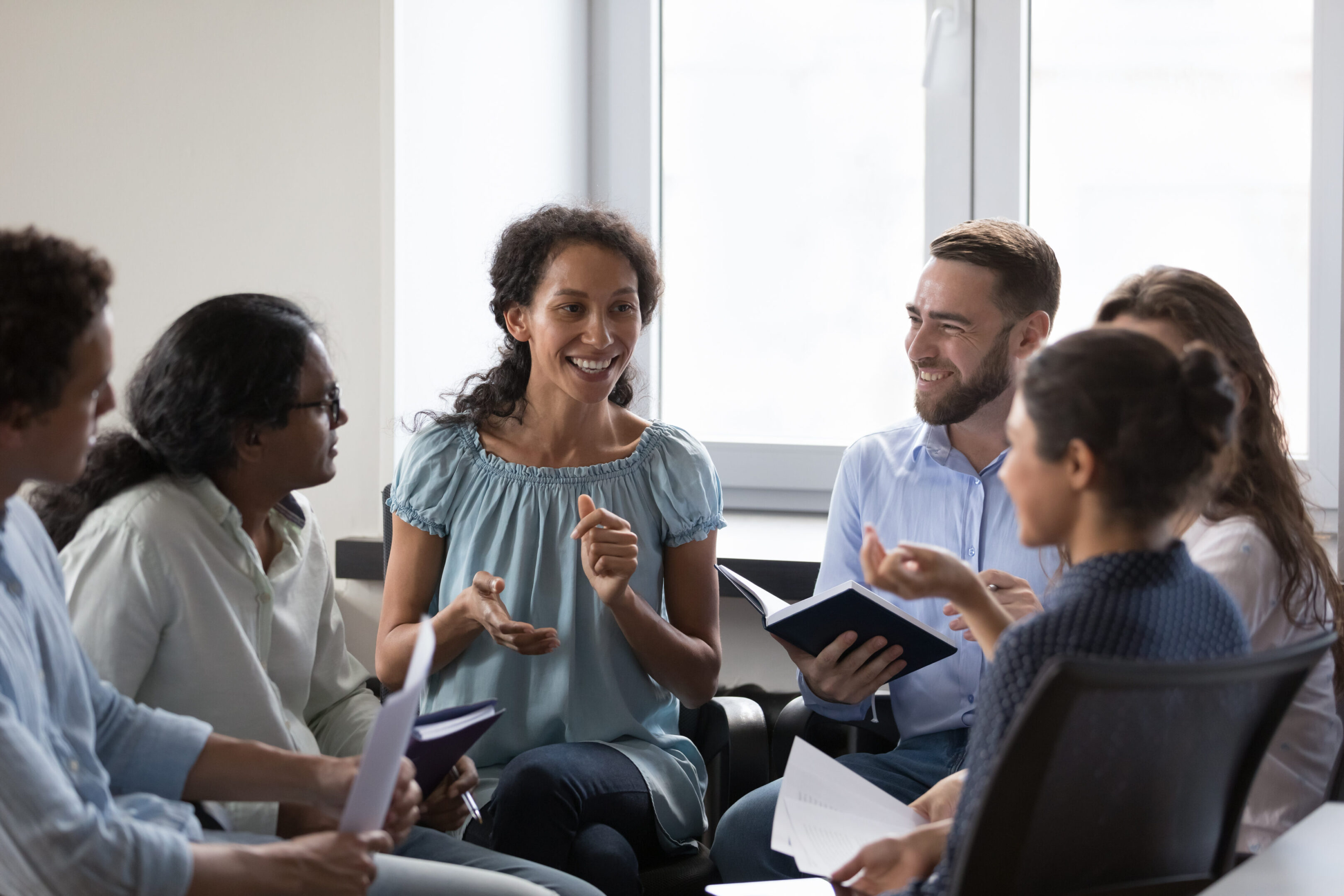 Happy diverse addicts sitting on chairs in circle, talking on group therapy meeting, discussing addiction, mental health problems. Multiethnic employees brainstorming on team training